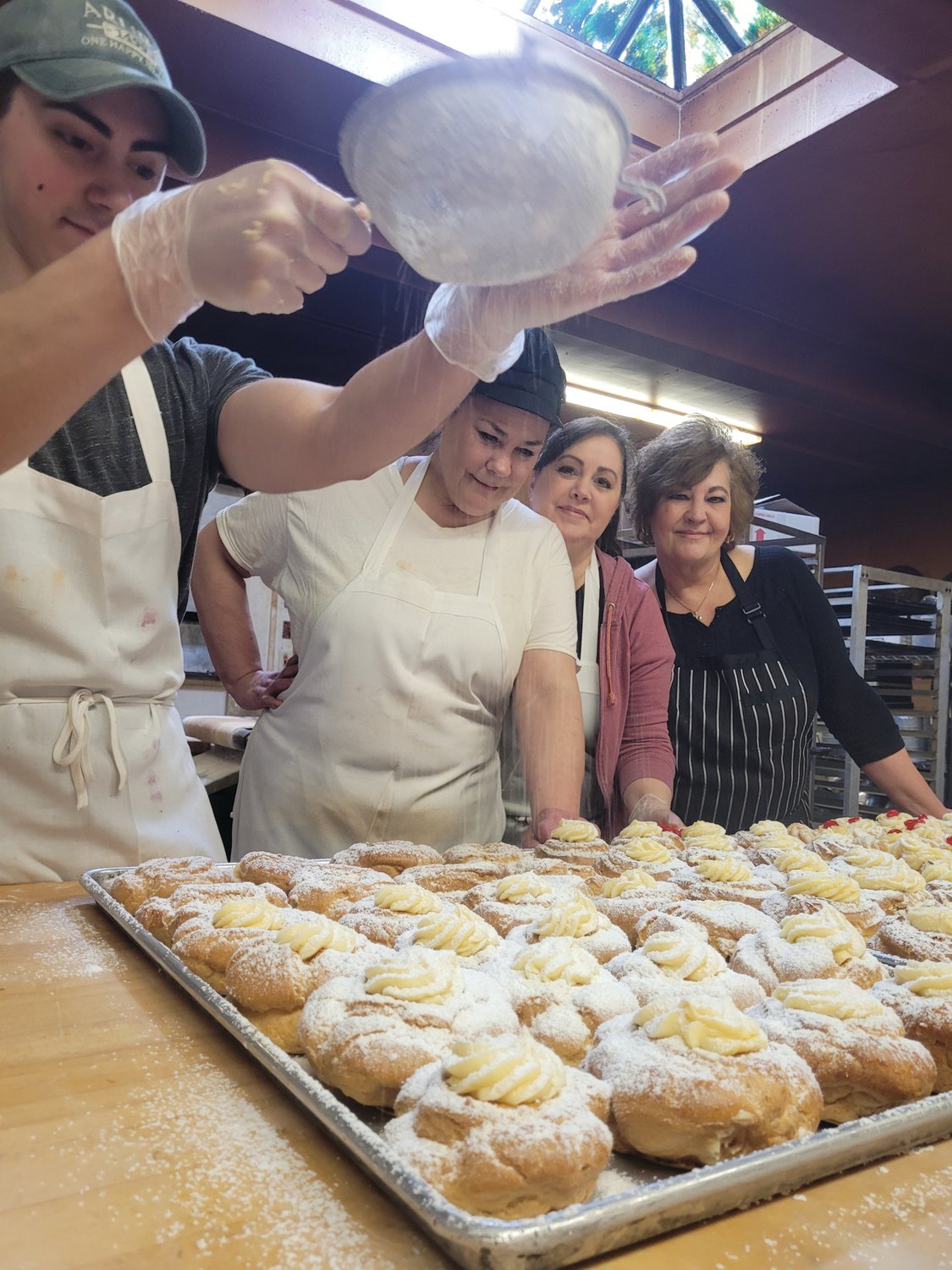 A DUSTING OF TRADITION: Brendan Notarianni shakes some sugar on a completed batch of zeppoles, as his aunt (and owner of Solitro’s Bakery) Elena Pennacchini, mother Diane Notarianni, and another aunt, Paula Arlia, watch on.